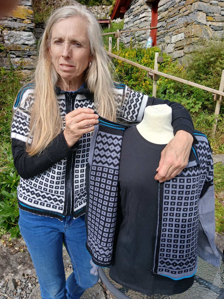Woman with long gray hair holding up a patterned knit vest outdoors, showing the navy and white geometric design