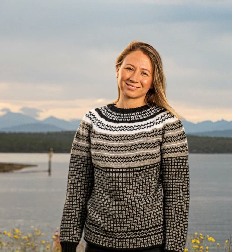 Woman in patterned sweater smiling outdoors with lake and mountains in background